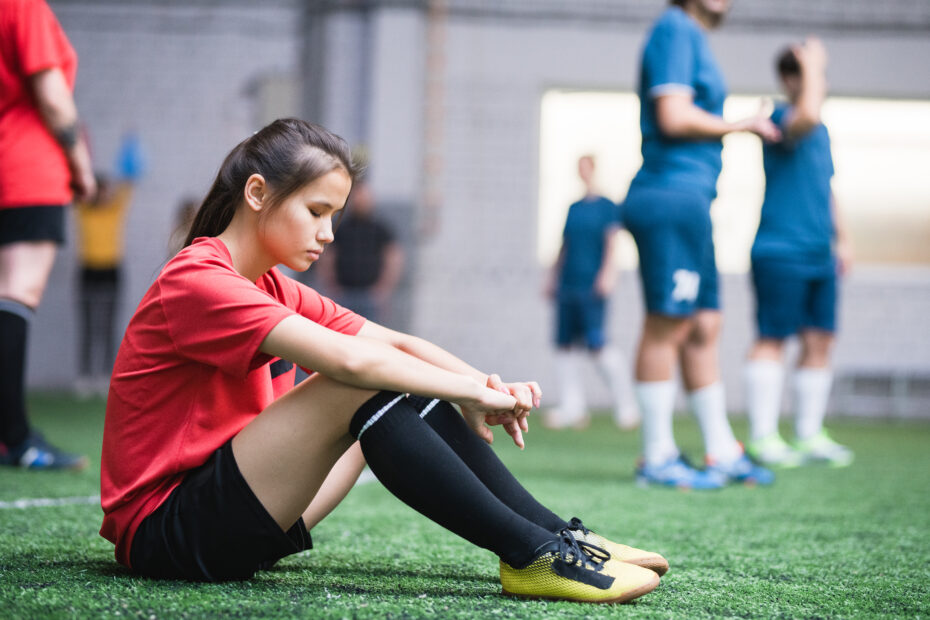 Sad or tired female football player in sports uniform sitting on green field